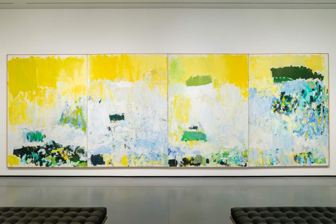 In a museum gallery hangs Salut Tom, a large abstract quadriptych painting. Yellow vertical marks make a sort of sky, and stippled white, green, and blue marks gather in a central, horizontal form.
