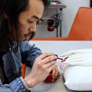 Christian Việt Đinh paints red nail polish onto ceramic hands with long fingernails. He is , a Vietnamese American man with long dark hair and a mustache wearing a denim jacket.