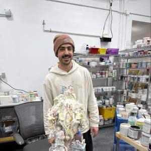 Sharif Farrag stands in a studio smiling in front of a detailed ceramic sculpture, surrounded by shelves full of ceramic glazes. He wears a brown beanie, beige hoodie, and has a medium light skintone, and a dark mustache.