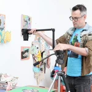 Thomas Friel, a white man with light skin tone, black and grey short hair, and glasses, adjusts a camera on a tripod in an art studio