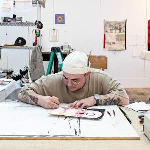 E Marshall draws on paper in a studio full of materials and artwork. He has a light skin tone, dark stubble beard, and wears round glasses, white beanie and has tattooed arms.