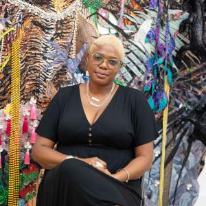 Ebony G. Patterson, a Black woman with medium dark skin, and short blonde curly hair, sits in front of a textile installation. She wears chunky acrylic glasses, a black dress and gold jewelry.