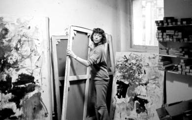 In a black and white photo, Joan Mitchell lifts a large canvas from a stack of paintings facing the wall by its stretchers, turning toward the camera. She is a white woman with dark hair in a bob, and wears a sweater with sleeves pushed up to her elbows and a wristwatch. On either side or her are vertical abstract paintings. To the right is a square window and a shelf holding boxes of tubes of paint.