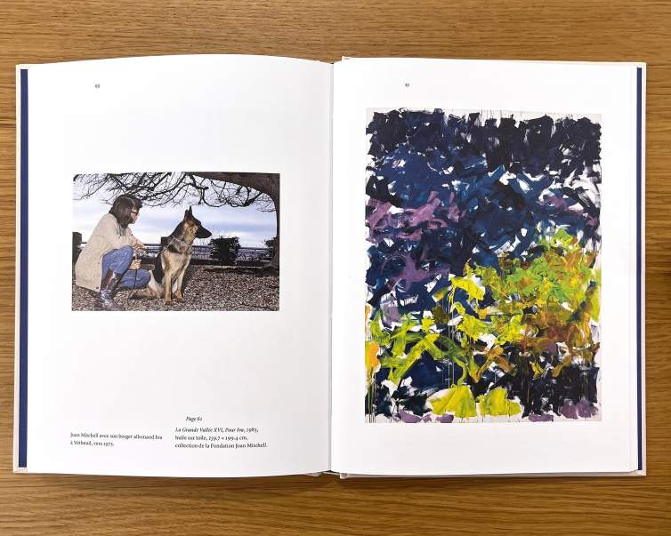 An open book on a wooden table shows a photo of the painter Joan Mitchell with a German shepherd on the left and a vibrant abstract painting on the right.