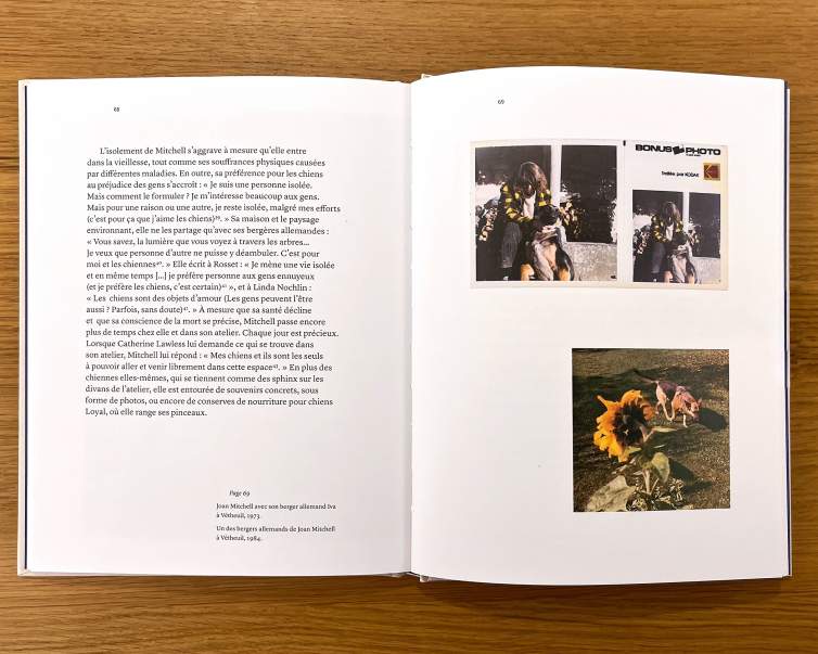 An open book on a wooden table shows a page of text and a page or archival photos of the painter Joan Mitchell with her dogs.