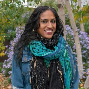 Chitra Ganesh smiles broadly in front of greenery, wearing a teal patterned scarf, denim jacket, gold nose rings and earrings. She has medium-dark skin, and long wavy dark hair.