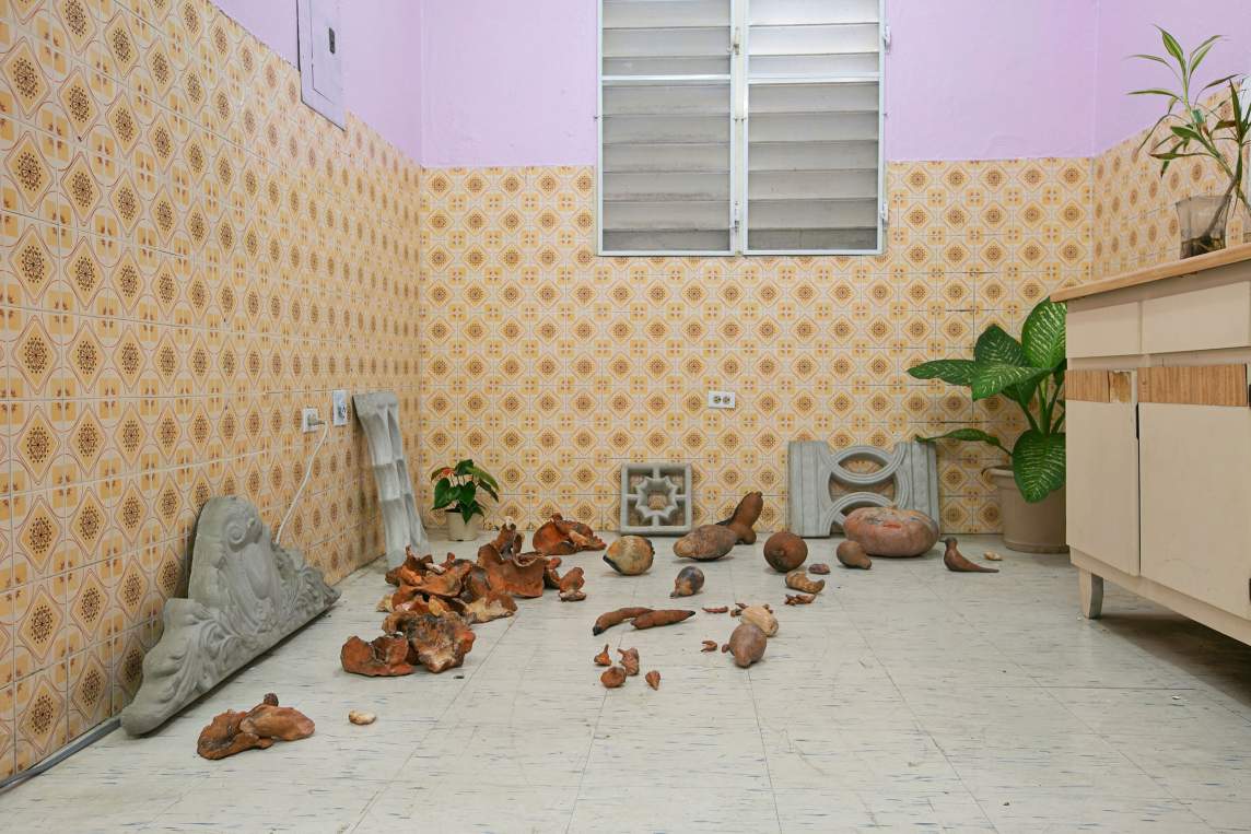 “Diamante Azul (Blue Diamond)” is an installation in a room lined with patterned yellow-brown tiles below lavender walls and a louvered window. Concrete balusters and decorative blocks lean against the wall, and around the linoleum floor are various brown and rust colored organic forms, resembling gourds, fruits, and tubers. A potted plant sits in the corner, next to a tan and wood vanity.