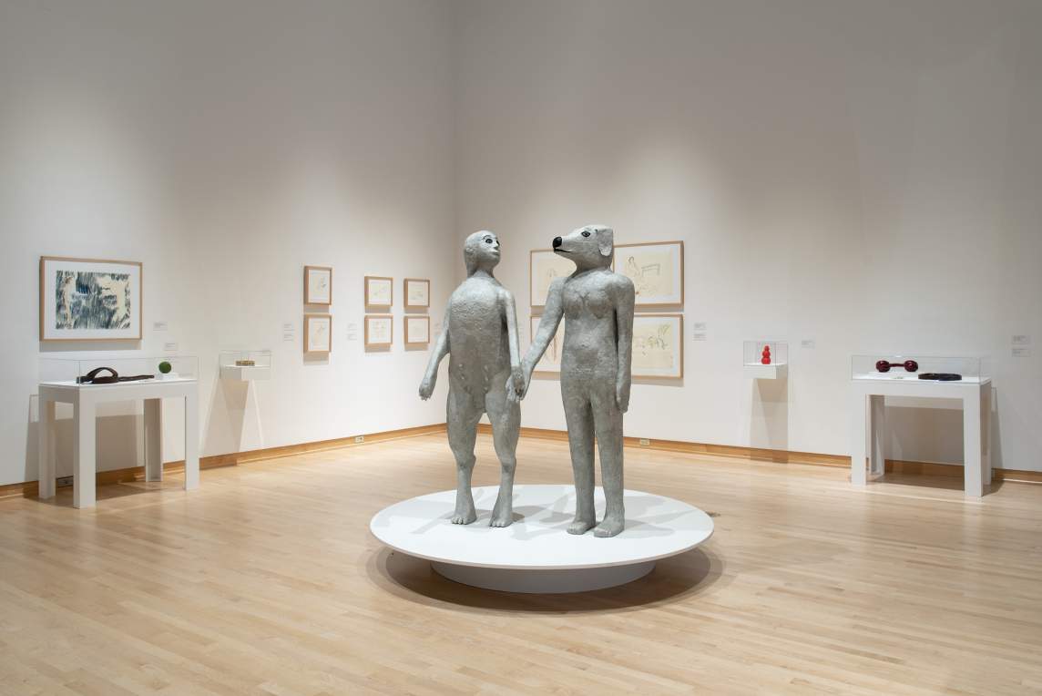 Two sculptures depicting hybrid human-animal figures stand side by side in a gallery space. They hold hands, with their heads slightly turned to gaze affectionately at one another. On the right, is a naked woman with a dog’s head; the figure on the left is an anthropomorphized dog with a long tail standing on its hind legs with the head of a woman.