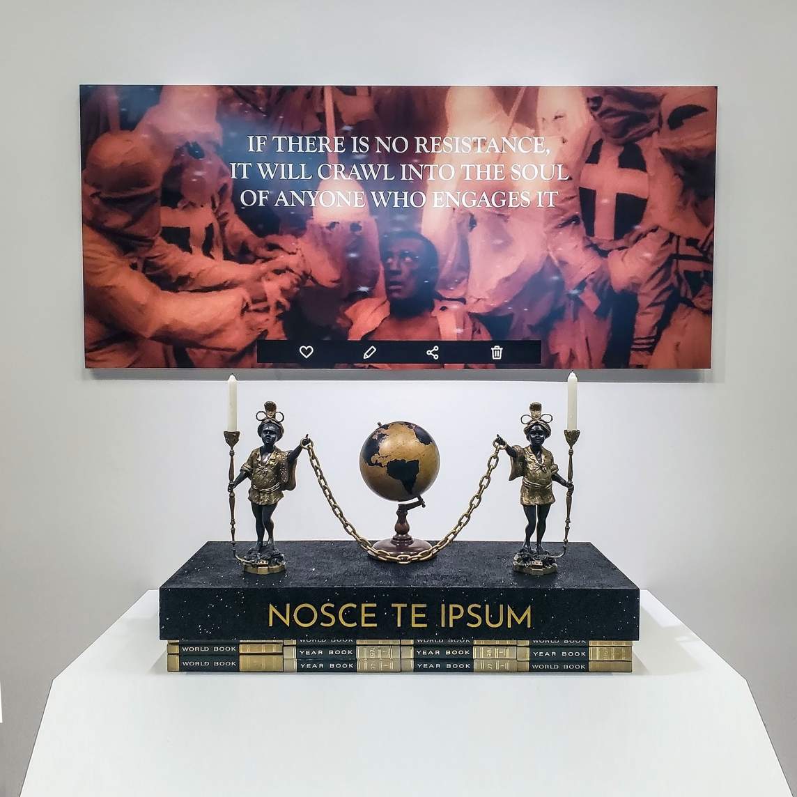 In the absence of light, flip the script in your mind is a sculptural installation. In the foreground are two metal figures holding candles in one hand and holding a chain between them, in front of a small globe. They stand on a platform reading “NOSCE TE IPSUM” stacked on top of books labeled World Book and Year Book. Behind this assemblage is  a sepia toned photo of a Black man surrounded by people in Klan robes with text overlaid “IF THERE IS NO RESISTANCE, IT WILL CRAWL INTO THE SOUL OF ANYONE WHO ENGAGES IT” and social media icons overlaid at the bottom.