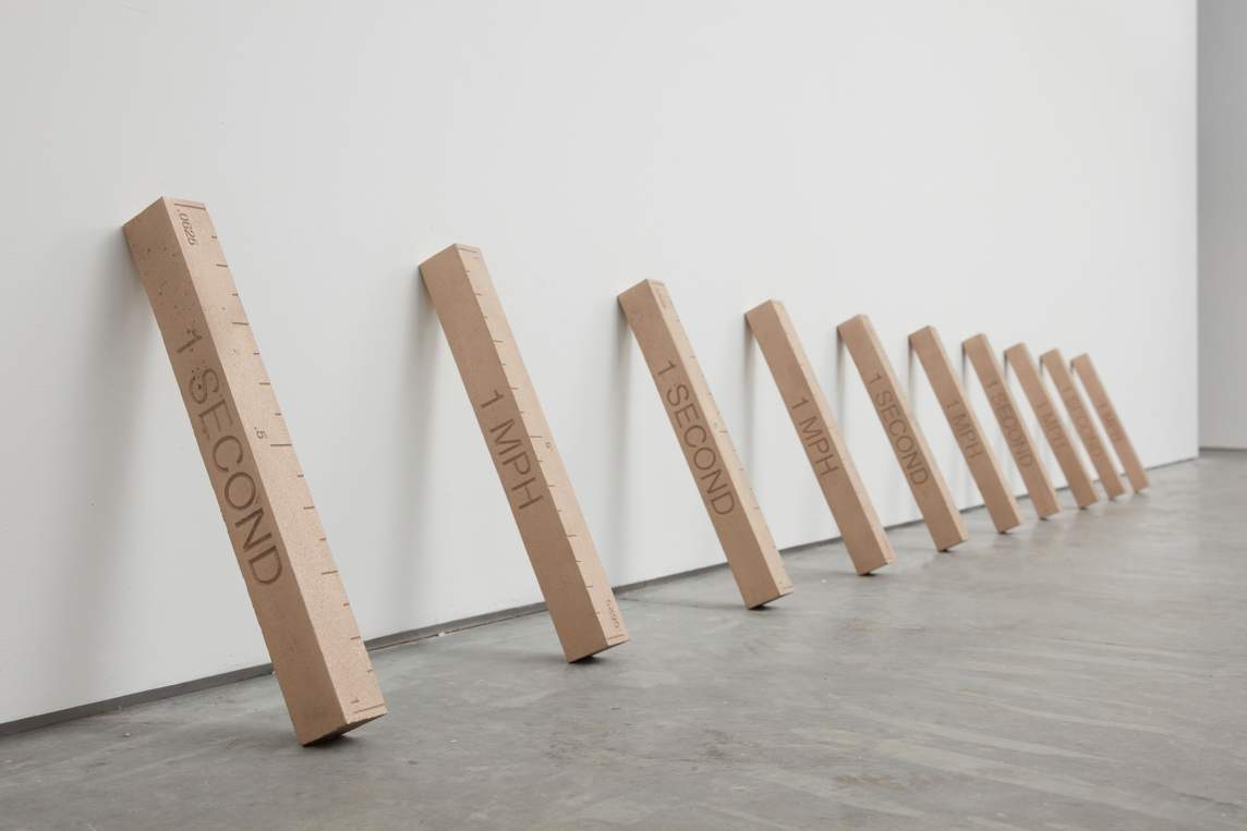 00:10 (To Remember the Murder of Eric Garner) is a row of 10 rectangular bronze columns leaning against a white wall reading alternately 1 SECOND and 1 MPH, with ruler measurements on the other face of the column.