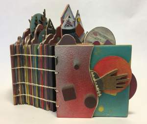 Jupiter in Our Sky is a sculptural book, with thick multicolor pages bound in a horizontal stitch. Fragments of buildings and shapes extend beyond the edges of the book, and the cover has a dimensional hand over a sphere.