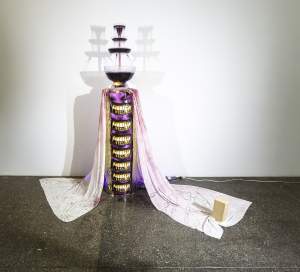 wtf is juice / gw smile is a plastic party fountain filled with cascading purple liquid operates on top of a column formed by a stack of images of President Washington's dentures with yellowed teeth. White velvet stained purple in places flows down from the top of the column, with a tan brick resting on the edge of the fabric.