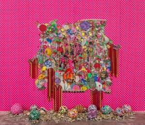 they were beautiful like the moon… is an intricately embellished textile with floral motifs and two figures at the center. Swags of beads drip down from sections and the piece hangs on a beaded polka dot wall. On the ground are balloons covered with patterns and a pile of gold plastic trophies.