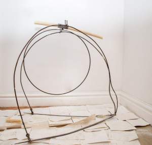 Semash is an abstract sculpture in the corner of a white room. On a bed of off-white paper sheets a loop of steel forms a loose arc and circle, bound by hardware at the apex which also holds a horizontal candle dripping wax onto the form.