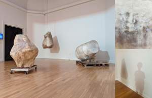 Subsiste is a sculptural installation in a white gallery room - 3 large objects are shaped like faceted boulders or bone fragments. One is on a shipping pallet, one on a rolling dolly, and the third is suspended from the ceiling by a chain.