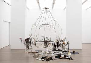 Black noodles is a large sculpture in a white space, centered on a large steel  chandelier hanging near the floor. Loops and tangles of string and rope hang from its arms, and on the floor, various objects are arranged on circles.