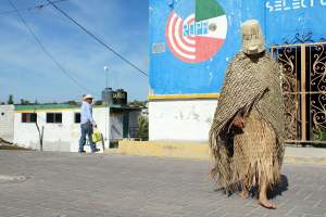 Kajne: Untitled II is an image of a performance where a barefoot figure walks through a public area, wearing a basket helmet obscuring their face, and wrapped in a cape and skirt woven from palms.