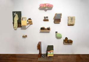 I Can Hear Your Dirt is a sculptural installation on white wall, of sculpted domestic items on shelves and brackets at varying scale, including a miniature table and chair, miniature tile wall and bathroom sink, corner of a sink with razor, slightly small work boots, small handing pants, and a ceiling surface with pink insulation and a fire detector.