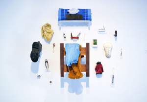 Custodian of The Ephemeral is a sculptural installation on a white wall, of various sculpted objects hung on the wall or standing on shelves. Objects include a work boot with the sole out, a spoon, swiss army knife, miniature hanging jacket, rolled blanket with sheep illustration, flower, and clothes hung over a detached bed frame.