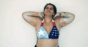 To be a disaster, to be diaspora, to be a state, to be an island, to be Puerto Rican is a film still of a woman wearing a bikini top decorated Puerto Rico and US flag motifs. She stretches her elbows up with her hands behind her head with eyes closed. Her arms and hands are smeared with white paint.