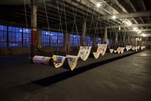 Art installation in an industrial space featuring large hanging scrolls with colorful images, suspended from the ceiling by wires in several rows, with dim lighting and visible windows in the background.