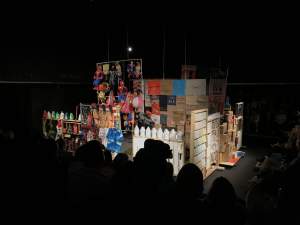 El maestro 2 is an assemblage installation in a darkened space of various objects arranged in vertical plinth on a stage-like area. Groups of items include handing marionettes, flattened paper shopping bags, rows of plastic bottles, shipping boxes, tourist towels and shirts and more.