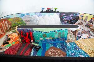 This detail of Snake Tube Adventure Racing shows the interior of a soft racetrack sculpture decorated with brightly colored quilts of landscapes scenes, and two toy vehicles with iPhones attached to their fronts are in the track.