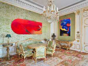 A lavish room with ornate furniture, a chandelier, and patterned wallpaper. Modern red and blue paintings decorate the walls, blending classical and contemporary design elements.