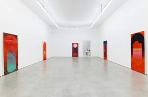 A brightly lit art gallery room features six framed abstract paintings mounted on white walls. The floor is concrete, and the room appears minimalist with a doorway at the back center.