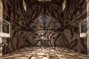 The Greys in Between consists of two intricately patterned geometric metal sculptures hang from the ceiling, casting complex shadow designs on the walls and floor of a gallery room.