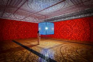 All the Flowers Are For Me (Turquoise) is a blue, ornate illuminated cube casting intricate patterns on the red walls and wooden floor of a gallery room.