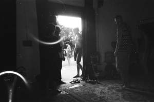 In a black and white photo, a group of people socialize near an open doorway, with some standing inside a dimly lit room and others outside. A rug and scattered shoes are visible on the floor.