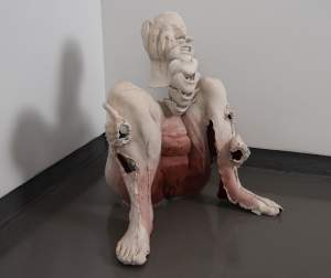 Agony is a figural sculpture of a naked and fragmented seated woman holding their knees. A wincing face has repeated chins stacked like a spine, and red pigment radiates from genitalia.