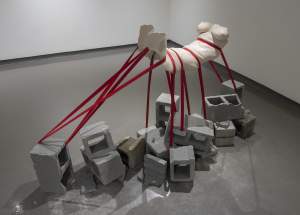 Weighted is a white female torso sculpture suspended above scattered concrete blocks by multiple red straps pressing into the flesh, in a gallery space with gray floors and white walls.