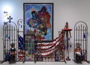 A mixed-media art installation featuring a framed painting, U.S. flag elements, metal gates, and sculptures. Blue bottles and a cross are also included in the display.