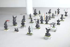 Campo Santo is an array of 20+ small ceramic sculptures on small grassy pedestals, arranged in a loose grid, of figurative forms that combine religious figurines with popular culture and personal mementos. Busts of people have animal ears, horns or small hats.