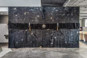 Before We Blast Off is a horizontal Intricate mural with geometric patterns, featuring a central clock surrounded by objects including hurricane lamps, tassels and netting, displayed on a dark wall in a spacious room.