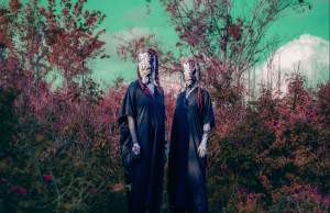 We’ve Been Wearing Your Skin We Have Felt How It Traps You shows two people in black robes and white, wrinkly textured face masks with no eyes stand side by side in a field of pinkish foliage with green sky and trees in the background.