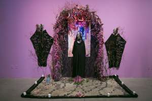 We've Been Among You is an art installation featuring a figure at the center draped in black with a textured mask, surrounded by an arch of branches and foliage, set against a purple wall with stones and candles on the floor. Flanked on each side are robes pinned to the wall with masks and handwritten words.