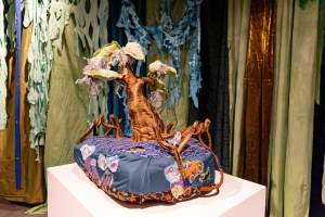 Fabric sculpture of a tree on a cushioned base, displayed on a pedestal. The background features hanging fabric in shades of green and blue.