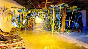 Art installation with large, colorful leaves and stems hanging from the ceiling in a gallery. The floor is reflective, and there are fabric elements arranged around the space.