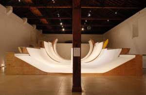 In a darkened gallery space, Number 03-16 (A FANAL) is a large symmetrical sculpture with wooden sides and a white top featuring 5 various curved surfaces for skateboarding.