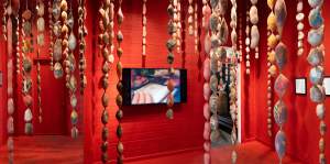 Art installation with hanging pantyhose filled like sausages in a red room, featuring a screen displaying images on the wall.