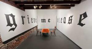 Gallery room with "grieve me please" in Gothic font on walls. Centered, an orange bathtub filled with flowers and flanked by two wooden chairs. Exposed beams on ceiling.