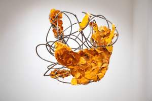 Abstract sculpture with intertwined metal rods and orange leaf-like elements, suspended in a white gallery space.