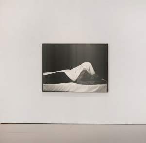 Framed black and white photo of a person lying down, displayed on a white gallery wall.
