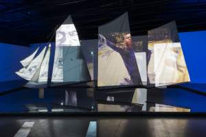 Art installation with sails displaying projected images, including close-up faces and a person with outstretched arms, set against a blue backdrop.