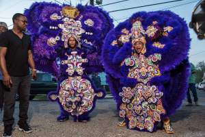 Two people stand in ornately decorated Indian masking suit, with detailed beaded medallions flanked by radiating plumage of purple feathers.