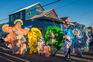 In front of a blue painted house a group of adults and children wear ornately  decorated beaded and feathered Indian masking suits in a rainbow of colors.