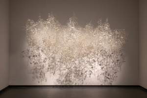 Whisper #2 is a suspended abstract sculpture made of thin, interwoven metallic wires  with oblong paper shapes, which casts intricate shadows on a white gallery wall and floor.